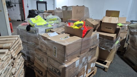 Warehouse storage area with stacked cardboard boxes, pallets, and neon safety vests organized on and around wooden crates