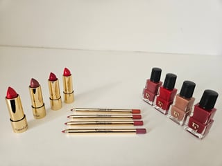 Collection of makeup products including gold lipsticks in red shades, matching nail polish bottles, and burgundy lip liners arranged on a white surface.