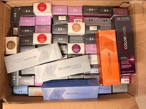 Cardboard box filled with various Revlon cosmetics and hair care products including makeup compacts, color bottles, and styling tools