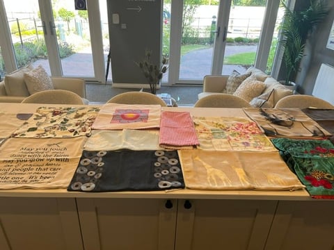 Wooden table displaying various fabric swatches and textile samples in different colors and patterns, with windows and chairs visible in the background