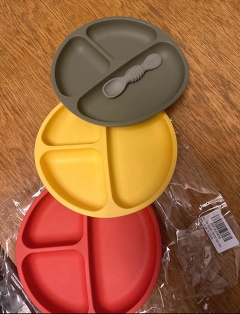 Three stacked silicone baby plates in gray, yellow, and red on a wooden surface, with a gray spoon on top