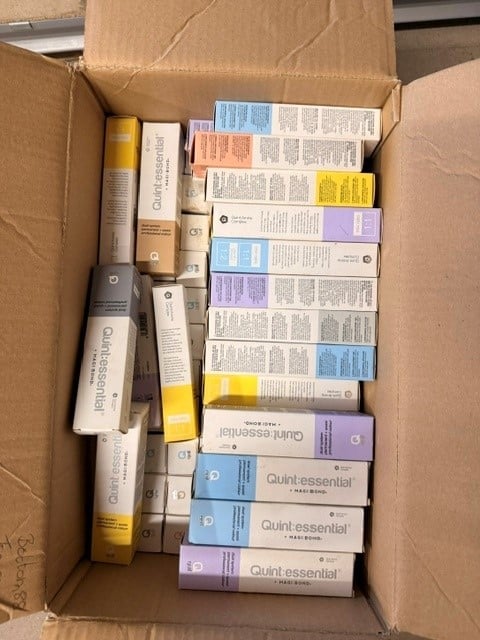 Cardboard box containing various beauty and skincare product boxes with colorful labels including Quint-essential and other cosmetic items