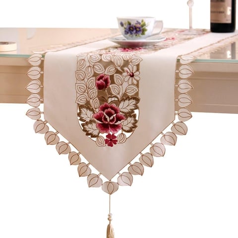 Beige table runner with embroidered red floral design and decorative scalloped edges with beaded trim and tassel, displayed on a dining table
